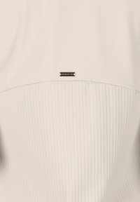 Beige Sportoberteil mit gerippter Textur im unteren Bereich, glattem oberen Bereich und einem schwarzen Logo-Label in der Mitte.