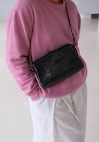 Borsa a tracolla nera in ecopelle con texture a coccodrillo, dotata di chiusura a zip e tracolla staccabile. Indossata sopra un maglione rosa.