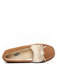 Zapatilla de ante marrón con forro de piel de oveja crema, costura decorativa y un lazo en la parte delantera. La suela es plana y texturizada para mayor agarre.