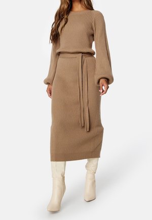 Femme portant une robe midi ceinturée en tricot côtelé beige avec des manches bouffantes, associée à des bottes blanches jusqu'aux genoux, se tenant devant un fond blanc.