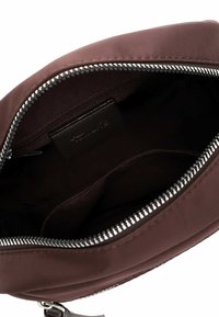 Atvērta brūna ādas soma ar rāvējslēdzēju, kurā redzama tumša iekšpuse ar mazu iekšējo rāvējslēdzēja kabatu un iekšā iešūtu etiķeti.