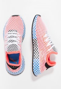 Baskets Adidas avec un design en maille, présentant des couleurs blanche, rouge et bleue. Semelle et accents noirs, avec une tige en textile.