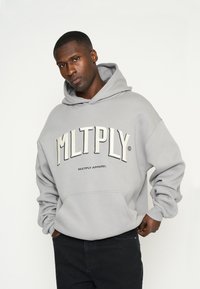 Grijze hoodie met een voorste kangoeroezak, met een opvallend geborduurd logo "MLTPLY" in het wit, tegen een gestructureerde stofachtergrond.
