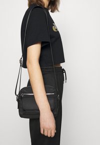 Černá crossbody taška se dvěma zipovými přihrádkami, hladkým povrchem a nastavitelným popruhem, držena vedle osoby v zkrácené černé košili.