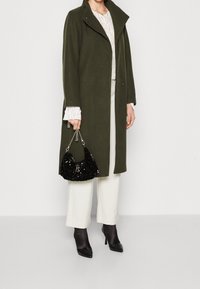 Olive green wool coat with a high collar, paired with a white lace top, cream trousers, black heeled boots, and a black sequined handbag.