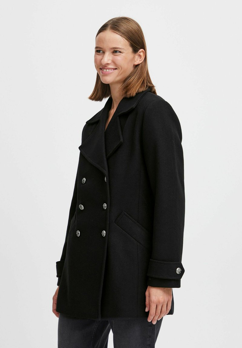 Manteau en laine noir avec un design croisé, grand col, quincaillerie en boutons argentés et deux poches latérales. Tissu texturé avec une coupe structurée.