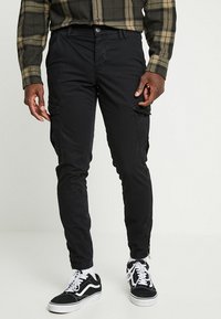 Man med svarta smala cargo-byxor, svarta och vita sneakers samt en grön och svart rutad skjorta som står mot en enfärgad bakgrund.