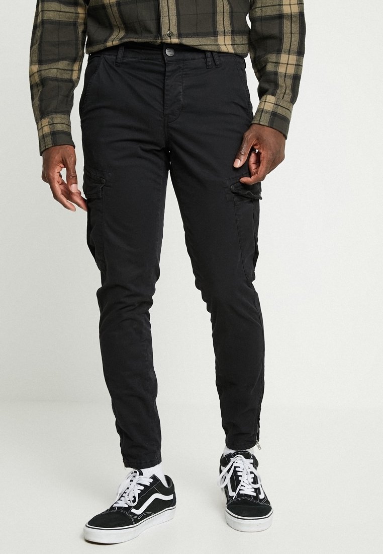 Man med svarta smala cargo-byxor, svarta och vita sneakers samt en grön och svart rutad skjorta som står mot en enfärgad bakgrund.