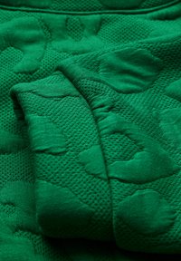 Grüner Strickstoff mit einem strukturierten Muster, das erhabene, abstrakte Formen zeigt. Das Material ist weich, mit einem gleichmäßigen Gewebe und einer glatten Oberfläche.