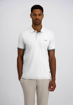 Homme portant un polo blanc à manches courtes avec un liseré noir sur les manches et un pantalon beige clair, debout devant un fond gris uni.