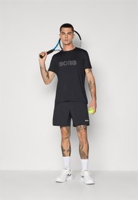 Svart treningstrøye med "BORG"-logo, paret med matchende shorts, hvite sportssko, og holder en tenniskølle og ball.
