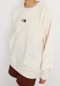 Sweat-shirt beige clair avec col rond et coupe décontractée. Présente un petit logo noir centré sur la poitrine. Texture lisse, manches longues.