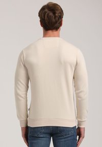 Sudadera de color beige claro con una textura suave, puños y dobladillo acanalados, y un pequeño parche en la parte posterior cerca del cuello. Llevada con jeans azules.
