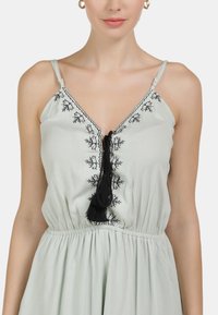 Robe vert clair avec des bretelles fines, broderie florale noire, pompon décoratif au niveau du décolleté et taille froncée pour la forme.