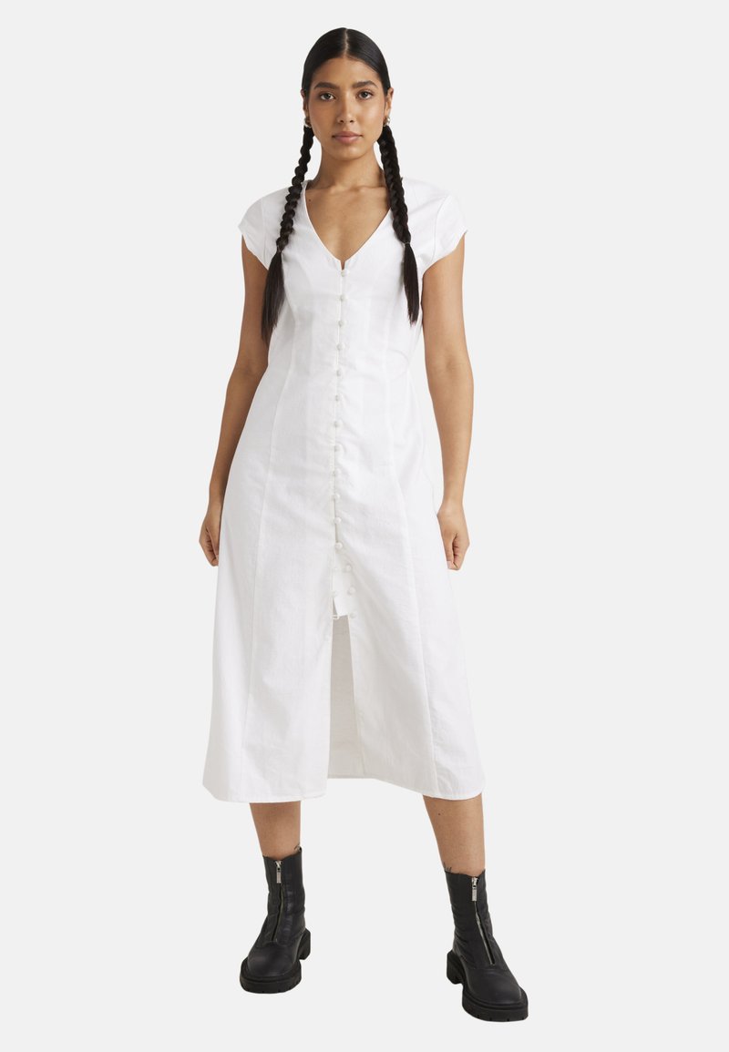Hvit midi-kjole med korte cap-ermer, V-hals, knappestolpe foran og splitt. Brukt med svarte ankelstøvler. Glatt tekstur, minimalistisk design.
