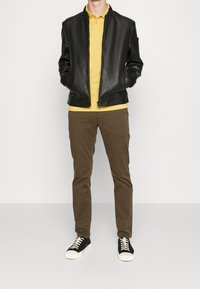 Černá kožená bomber bunda přes žluté polo tričko, doplněná hnědými chinos kalhotami a černými teniskami s bílými tkaničkami. Jednoduchý design a střih.
