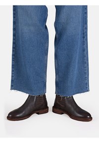 Bottines en cuir marron avec des panneaux latéraux élastiques, associées à un jean bleu à large jambe avec ourlet effiloché. Les semelles des bottines ont une texture avec une finition en bois.