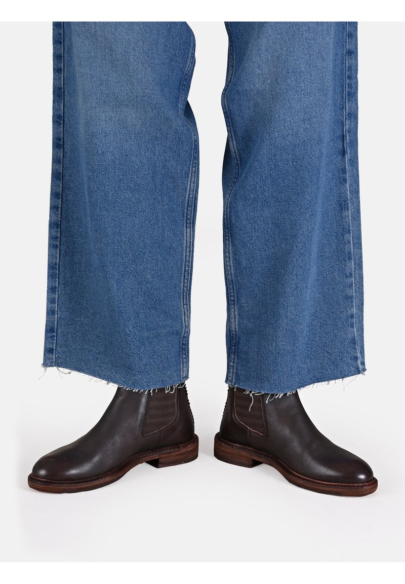 Bottines en cuir marron avec des panneaux latéraux élastiques, associées à un jean bleu à large jambe avec ourlet effiloché. Les semelles des bottines ont une texture avec une finition en bois.
