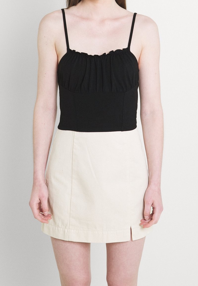 Femme portant un haut noir froncé sans manches et une mini-jupe beige avec une petite fente à l'avant, debout devant un fond uni.