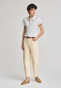 Vit kortärmad polo med fem knappar, kombinerad med ljust beige, löst sittande byxor och bruna loafers. Slät tygtextur.