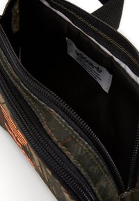 Černobílý a zelený maskáčový batoh vyrobený z odolného materiálu se zipovým uzávěrem, s oranžovým logem a přihrádkou s štítkem uvnitř.