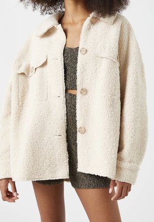 Personne portant une veste sherpa couleur crème par-dessus un crop top en tricot gris et un short assorti, debout devant un fond uni.