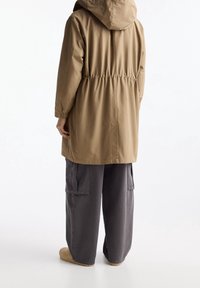 Parka à capuche kaki avec une taille élastique, des manches longues et une coupe décontractée, associée à un pantalon cargo gris. Tissu texturé.