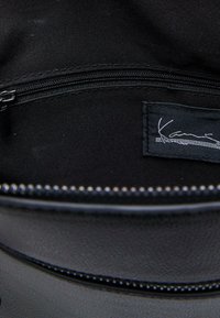 Unutrašnjost crne kožne torbe sa zatvorenim džepom. Ima glatku teksturu i tkanu oznaku s imenom "Kavi" u bijelom šavu.