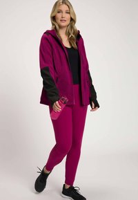 Sportoutfit met een fuchsia zip-up hoodie met zwarte accenten, fuchsia leggings en zwarte schoenen. Houdt een roze waterfles vast.