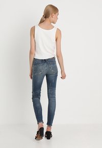 Jeans ajustados en denim azul deslavado con un acabado desgastado. Tiene cinco bolsillos, trabillas para cinturón y un diseño ajustado. Combinados con una camiseta sin mangas blanca.