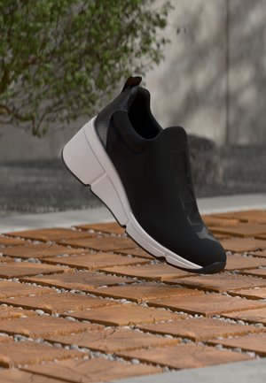 Čierna slip-on teniska s bielou podrážkou a vyrazeným logom „DKNY“, vznášajúca sa nad dreveným povrchom s rozptýlenými malými kameňmi.