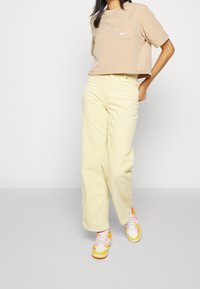 Person som bär en beige beskuren Nike t-shirt, ljusgula vida byxor och färgglada sneakers står framför en helt vit bakgrund.