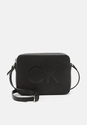 Czarna prostokątna torba na ramię z wytłoczonym logo "CK", regulowanym paskiem i zasuwanym zamknięciem na górze na jednolitym tle.