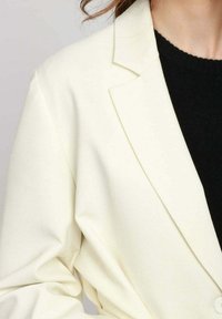 Blazer bianco con una vestibilità su misura, rever a scollo, cuciture dettagliate e chiusura con un solo bottone, indossato sopra un indumento nero.