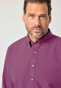 Bärtiger Mann mit kurzen lockigen Haaren, der ein violettes Hemd mit Knöpfen trägt, auf dessen Brust "JP 1880" gestickt ist, und geradeaus schaut.