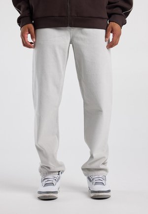 Person trägt hellgraue Jeans, weiße und graue Sneakers sowie eine dunkelbraune Jacke mit Reißverschluss und steht mit entspannten Händen an den Seiten.