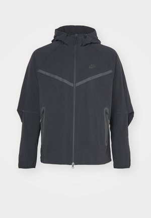 Veste Nike noire avec fermeture éclair sur le devant, capuche et coutures angulaires. Comprend des accents réfléchissants et des poches latérales zippées. Fabriquée en tissu léger.