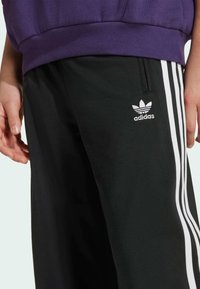 Pantalons de survêtement noirs Adidas avec des rayures blanches sur les côtés, fabriqués en tissu lisse. Présente un petit logo blanc sur la cuisse et un design à coupe décontractée.