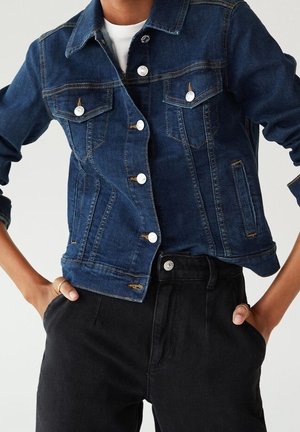 Personne portant une veste en denim bleu foncé sur une chemise blanche, les mains dans les poches d'un pantalon noir taille haute.