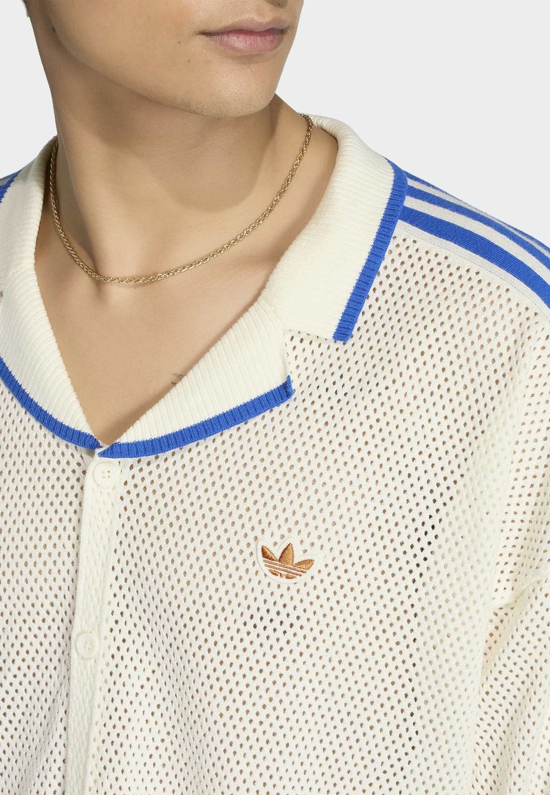 Jongere draagt een wit mesh poloshirt met blauwe bies en een klein bruin Adidas-logo, en een dunne gouden ketting.