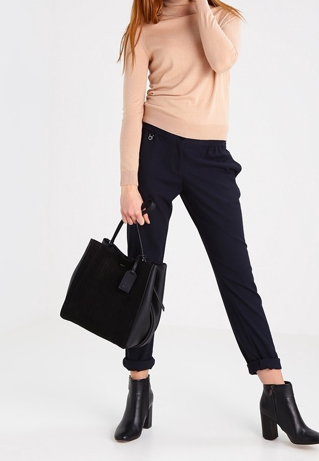 Černá kožená taška přes rameno s vertikálními žebrovanými texturami, horními uchy a odnímatelným štítkem. V kombinaci s námořnickými kalhotami a béžovým topem.