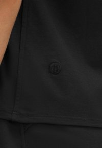 Svart atletisk topp med en slät yta, med en liten, ton-i-ton logobadge och rena sömmar längs fållen.