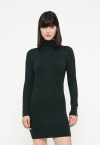 Mujer vestida con un vestido suéter ajustado de punto cableado en verde oscuro, con detalles de botones en los puños de las mangas, de pie frente a un fondo blanco.