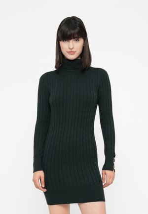 Donna che indossa un vestito maglione a collo alto attillato di colore verde scuro con dettagli di bottoni sui polsini delle maniche, in piedi contro uno sfondo bianco.