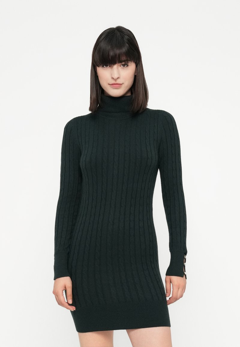 Mujer vestida con un vestido suéter ajustado de punto cableado en verde oscuro, con detalles de botones en los puños de las mangas, de pie frente a un fondo blanco.