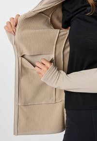 Une personne vêtue de noir et beige ouvre une veste beige côtelée pour révéler deux poches intérieures spacieuses.