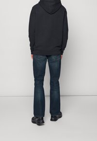 Sudadera con capucha negra, jeans de mezclilla azul de lavado oscuro, con perneras acampanadas, costuras visibles y zapatillas negras. Un atuendo sencillo y casual visto desde atrás.