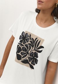 Camiseta de algodón blanca con un diseño floral negro sobre un fondo beige, de manga corta y con cuello redondo.