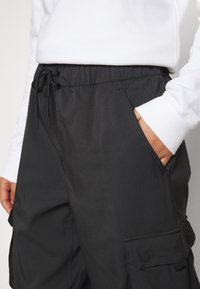 Person iført sorte cargobukser med sidelommer og en hvid langærmet top, med den ene hånd i lommen.