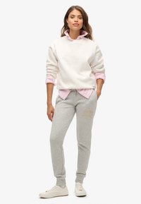 Biała, oversize'owa bluza nałożona na jasnoróżową koszulę z kołnierzykiem, zestawiona z jasnoszarymi dresami z złotym nadrukiem oraz białymi sneakersami.